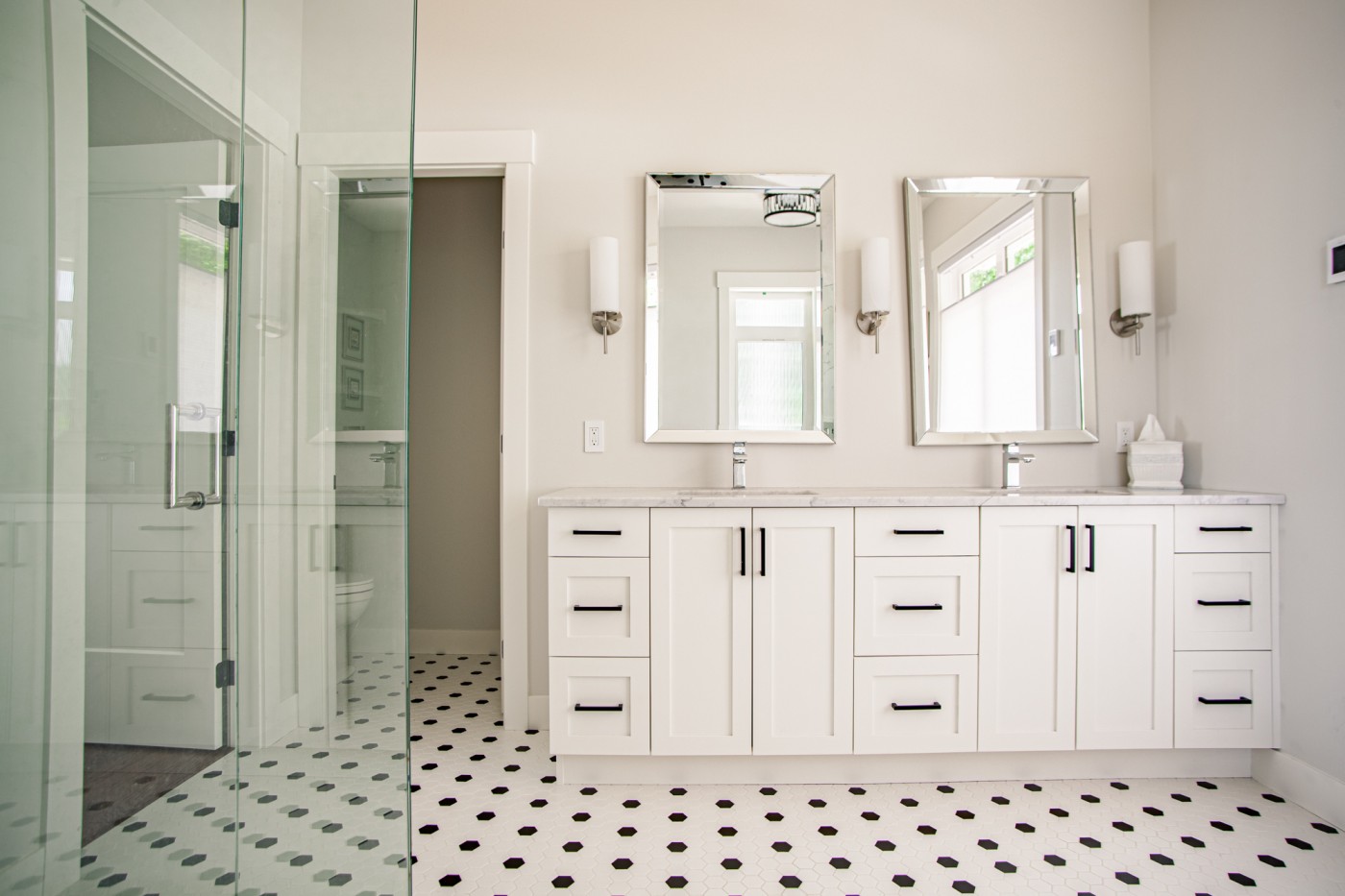 Bright white bathroom featuring a shaker-style double vanity with black hardware, marble countertop, and dual framed mirrors, complemented by wall sconces and classic black-and-white hexagon tile flooring.