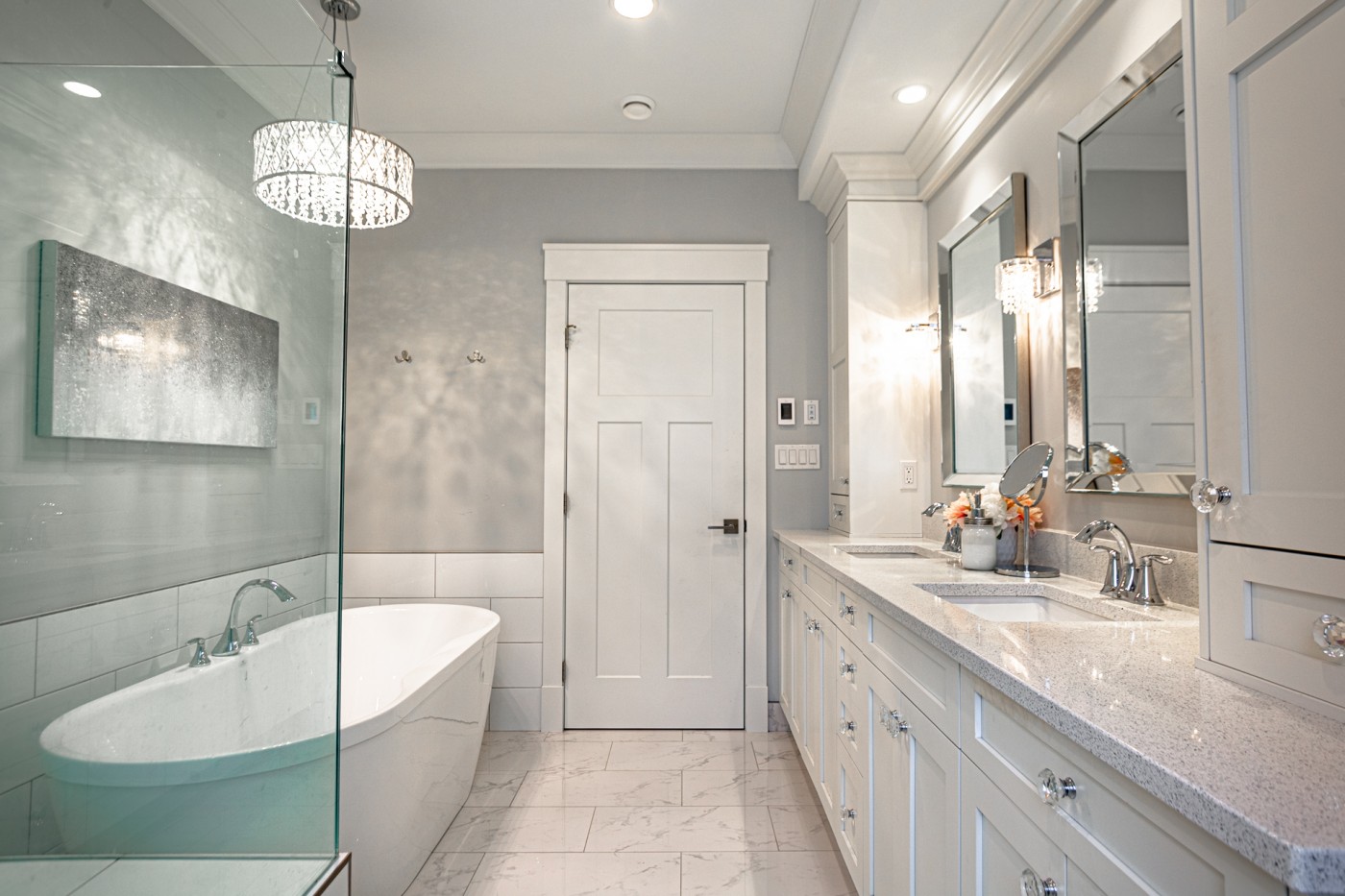 Elegant white bathroom with a freestanding tub, glass shower enclosure, and a long double vanity with shaker-style cabinetry, quartz countertops, and crystal hardware, accented by chrome fixtures, wall sconces, and a decorative chandelier.