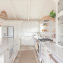 White boho kitchen with a quartz island, ceiling-height cabinetry, natural wood range hood, woven pendant light and champagne bronze hardware.