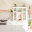 White boho kitchen with a custom wood-trimmed range hood, glass-front cabinetry, open shelves and potted plants.