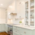 Two-tone traditional kitchen with soft green lower cabinets, warm white Merit upper cabinetry, glass display doors, subway tile backsplash, stainless steel range, pot filler, and brass hardware.