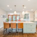 Bright traditional kitchen design with a soft green island, warm white perimeter cabinets, stainless steel range, leather island seating, hardwood floors, and patio doors.