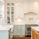 Bright traditional kitchen with soft green lower cabinets, warm white uppers, farmhouse sink, glass cabinet doors, subway tile backsplash, stainless steel range, and island seating.