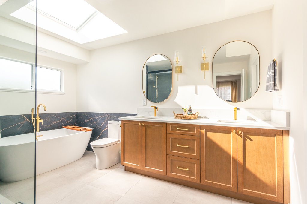 Bathroom renovations in Nanaimo example by Classic Kitchens & Design featuring a bright modern bathroom with natural light from a skylight, a freestanding white tub with gold fixtures, a wood vanity with shaker-style doors and brass hardware, white quartz countertop with dual sinks, and two round mirrors with gold sconces above.