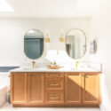 Double bathroom vanity with warm alder Lectus cabinetry, white quartz countertop, oval mirrors, gold faucets, matching wall sconces, and dark tile tub surround beside the vanity.