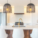 Symmetrical kitchen design with warm white cabinetry, rustic oak island panels, gold faucet, black woven pendant lights, stainless steel fridge, and centred custom range hood.