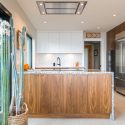 West coast modern kitchen with tall indoor plants beside the sliding glass doors, a walnut island, white cabinetry, quartz counters, built-in appliances, and warm wood storage.
