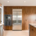Walnut kitchen storage wall with integrated stainless steel fridge, built-in wall oven, flat-panel pantry cabinetry, and quartz island countertop in the foreground
