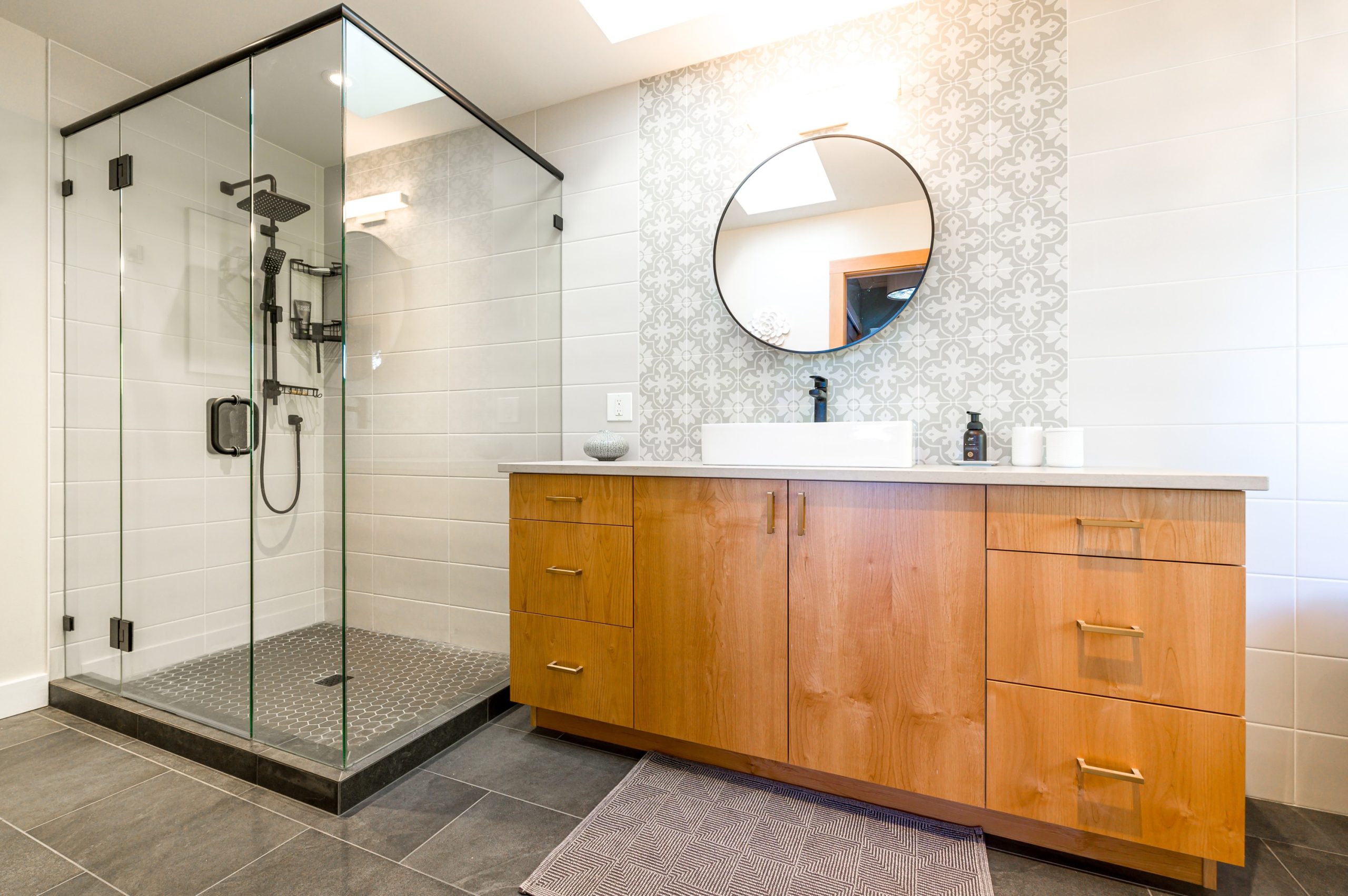 Bathroom renovations Nanaimo example by Classic Kitchens & Design showcasing a modern bathroom with a glass-enclosed shower featuring black fixtures, a floating wood vanity with brass handles and a gray countertop, patterned accent tile behind a round mirror, and dark gray floor tiles.