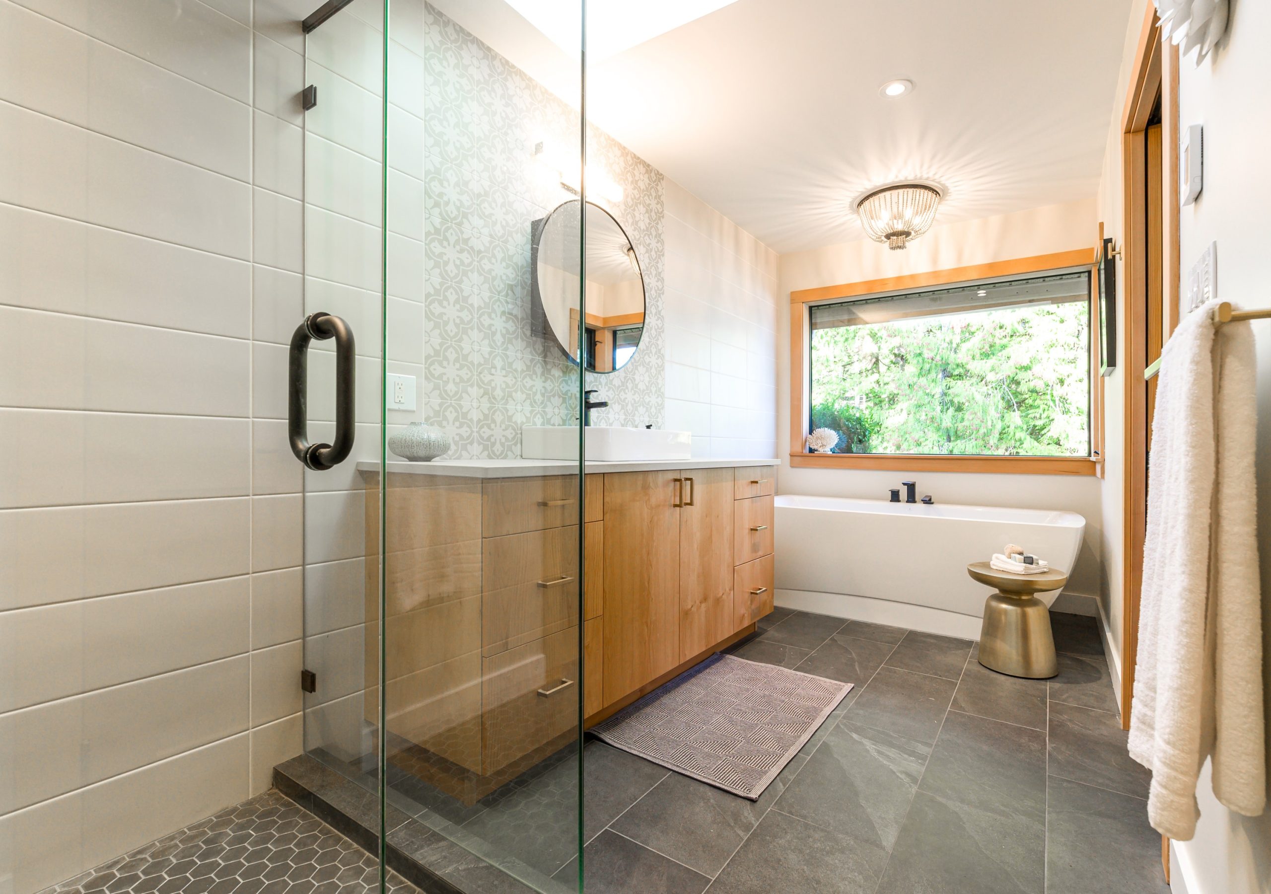 Modern bathroom with a glass walk-in shower, light wood vanity, and patterned feature wall, featuring a vessel sink, freestanding bathtub beneath a large window, dark tile flooring, and a gold side table for a touch of warmth and contrast.