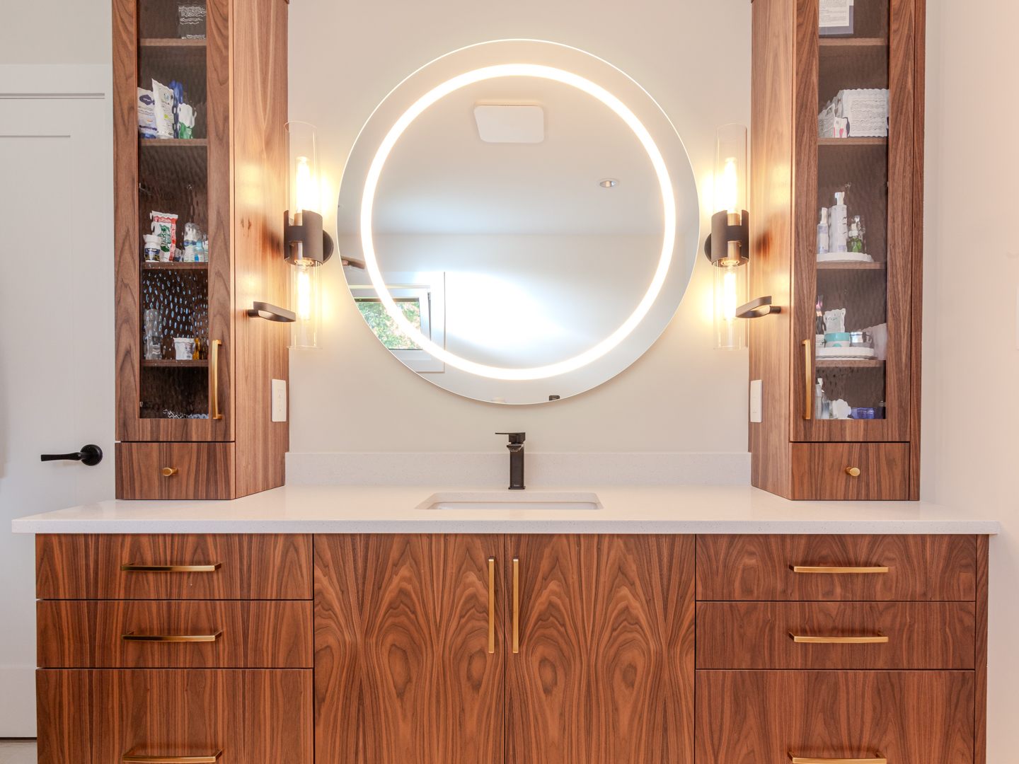 Bathroom renovation by Classic Kitchens & Design, featuring a contemporary bathroom vanity with rich walnut cabinetry, brass hardware, and a white quartz countertop,a round backlit mirror with modern wall sconces and glass-front tower cabinets on each side.