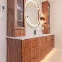 Natural walnut bathroom vanity with tall storage cabinets, illuminated round mirror, light countertop, brass hardware, and warm under-cabinet lighting