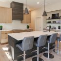 Kitchen island seating area with grey bar stools, Merit Kitchens maple slab cabinetry, white quartz countertop, dark island base, and warm wood flooring