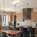 Open-concept kitchen and dining area with modern maple cabinetry, dark island, quartz countertops, custom range hood canopy, built-in appliances, and warm wood flooring